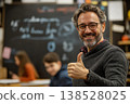 Smiling bearded man with glasses giving thumbs up in classroom, chalkboard with blurred writing behind him 138528025