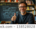 Smiling teacher in glasses giving thumbs up in classroom, chalkboard behind with blurred writing and bookshelf background 138528028