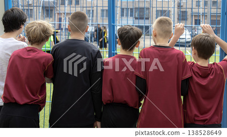 Back view of teenage boys in football uniforms watching a soccer match behind a blue fence Back view of teenage boys in football uniforms watching a soccer match behind a blue fence 138528649