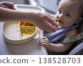 White Infant Hesitant To Accept Spoon, Parent Hand Reaching Across Tray, Messy Bib And Bright Orange Puree, Quiet Tension And Patient Encouragement In Home Feeding Scene 138528703