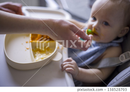 White Infant Hesitant To Accept Spoon, Parent Hand Reaching Across Tray, Messy Bib And Bright Orange Puree, Quiet Tension And Patient Encouragement In Home Feeding Scene White Infant Hesitant To Accept Spoon, Parent Hand Reaching Across Tray, Messy Bib And Bright Orange Puree, Quiet Tension And Patient Encouragement In Home Feeding Scene 138528703