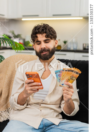 Indian man counting euro money cash salary using smartphone calculator for vacation budget future Indian man counting euro money cash salary using smartphone calculator for vacation budget future 138530377