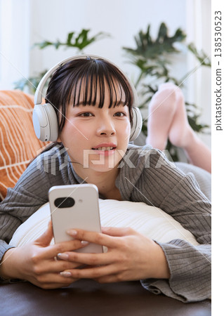 A woman enjoying her smartphone with headphones on; a relaxed lifestyle in the living room. A woman enjoying her smartphone with headphones on; a relaxed lifestyle in the living room. 138530523