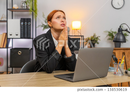 Young businesswoman folds hands praying before laptop looking up hoping project approval and order Young businesswoman folds hands praying before laptop looking up hoping project approval and order 138530619