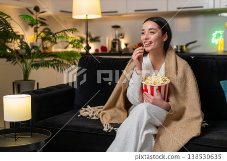 Young woman wrapped in plaid holds popcorn watches comedy on tv laughs comments comfy cozy evening Young woman wrapped in plaid holds popcorn watches comedy on tv laughs comments comfy cozy evening 138530635