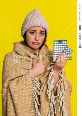 Woman wrapped in blanket wearing hat holding pills thoughtfully looking at them cold flu treatment Woman wrapped in blanket wearing hat holding pills thoughtfully looking at them cold flu treatment 138530722