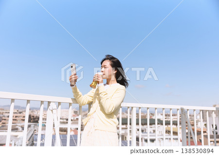 A woman enjoying a drink outdoors while holding her smartphone; a lifestyle of taking selfies under the blue sky. A woman enjoying a drink outdoors while holding her smartphone; a lifestyle of taking selfies under the blue sky. 138530894