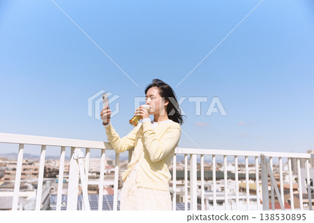 A woman enjoying a drink outdoors while holding her smartphone; a lifestyle of taking selfies under the blue sky. 138530895