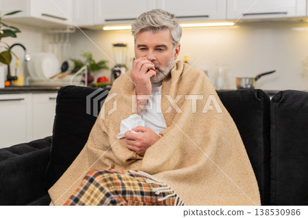 Middle-aged ill man under plaid sneezes uses tissue sniffles sick flu virus home quarantine rest Middle-aged ill man under plaid sneezes uses tissue sniffles sick flu virus home quarantine rest 138530986