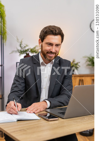 Middle-aged businessman attends webinar on laptop wears earphones takes notes snaps smartphone photo 138531090
