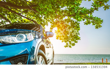 Blue sport SUV car parked by the tropical sea under umbrella tree. Summer vacation at the beach. Summer travel by car. Road trip. Automotive industry. Hybrid and electric car concept. Summer vibes. 138531394