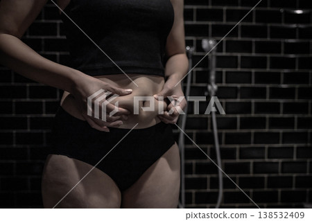 A young woman pinches the excess fat on her slightly chubby stomach. She's just starting to gain weight. She's in the bath. A young woman pinches the excess fat on her slightly chubby stomach. She's just starting to gain weight. She's in the bath. 138532409