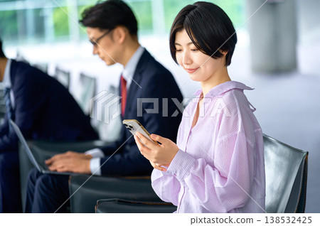 一位女士正在使用智慧型手機，一位商務人士正在辦公室休息室工作。 138532425