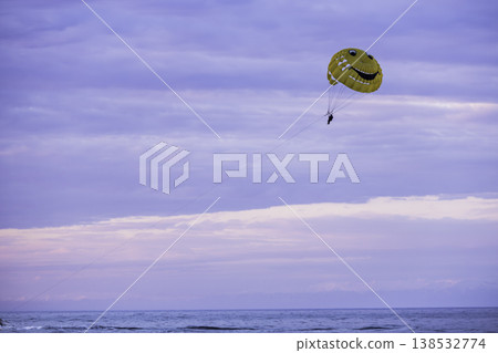 Parasailing with bright yellow smiley parachute in pastel sunset sky, lone person flying over sea horizon, minimal composition with soft gradient clouds, summer adventure and freedom concept 138532774