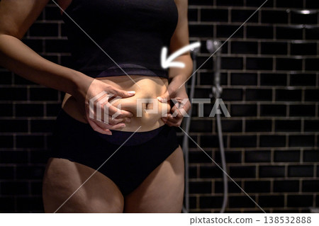 一位年輕女子捏了捏她略微豐滿的肚子上多餘的脂肪。她剛開始變胖。她正在洗澡。圖中用圓圈箭頭標示了她豐滿的肚子部位。 138532888