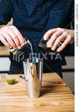 Hands pour blue and clear liquids from jigger and glass into stainless steel shaker. Craft cocktail movement, professional bartender, mixology techniques, artisan drinks, cocktail preparation. 138534142
