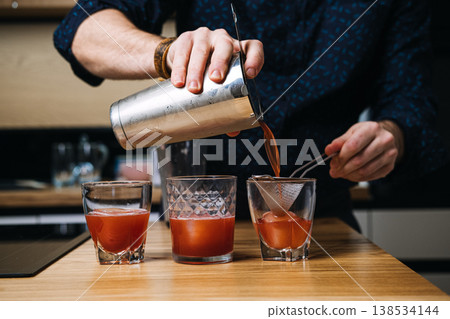 Bartender pours red liquid from metal shaker through strainer into glass with ice. Craft cocktail making, mixologist hands, beverage creation. Bartender pours red liquid from metal shaker through strainer into glass with ice. Craft cocktail making, mixologist hands, beverage creation. 138534144