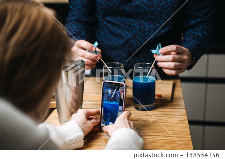 Hands hold smartphone to take picture of blue beverages in glasses on wooden table. cocktail masterclass, culinary education, group workshop, DIY drinks, food styling, behind the scenes. Hands hold smartphone to take picture of blue beverages in glasses on wooden table. cocktail masterclass, culinary education, group workshop, DIY drinks, food styling, behind the scenes. 138534156