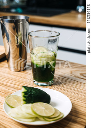 Mixing glass containing green liquid and cucumber slices sits on wooden counter next to metal shaker and lime. green juice cocktails, chlorophyll drinks, liquid wellness, vegetable-forward mixology Mixing glass containing green liquid and cucumber slices sits on wooden counter next to metal shaker and lime. green juice cocktails, chlorophyll drinks, liquid wellness, vegetable-forward mixology 138534158