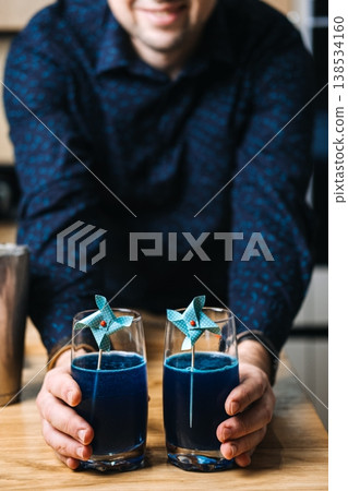 Man holds two glasses of blue liquid decorated with paper pinwheels on wooden table. cocktail masterclass, culinary education, group workshop, DIY drinks, food styling, behind the scenes. Man holds two glasses of blue liquid decorated with paper pinwheels on wooden table. cocktail masterclass, culinary education, group workshop, DIY drinks, food styling, behind the scenes. 138534160