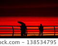 Silhouettes of people with umbrella on pier at red sunset over sea, dramatic sky with vivid orange and crimson tones, coastal lifestyle scene, evening walk by ocean horizon 138534795