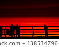 Silhouettes of people with umbrella on pier at red sunset over sea, dramatic sky with vivid orange and crimson tones, coastal lifestyle scene, evening walk by ocean horizon 138534796