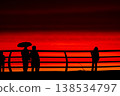 Silhouettes of people with umbrella on pier at red sunset over sea, dramatic sky with vivid orange and crimson tones, coastal lifestyle scene, evening walk by ocean horizon 138534797