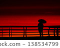 Silhouette of person with umbrella on pier at red sunset over sea, minimal coastal scene with dramatic sky and ocean horizon, evening mood with vivid orange and crimson tones 138534799