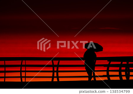 Silhouette of person with umbrella on pier at red sunset over sea, minimal coastal scene with dramatic sky and ocean horizon, evening mood with vivid orange and crimson tones Silhouette of person with umbrella on pier at red sunset over sea, minimal coastal scene with dramatic sky and ocean horizon, evening mood with vivid orange and crimson tones 138534799