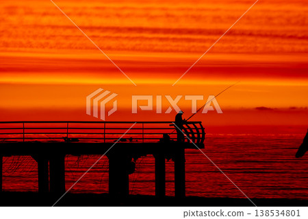 Silhouette of fisherman on pier at sunset over sea, dramatic red sky and ocean horizon, peaceful fishing scene, solitude and lifestyle concept with calm evening atmosphere Silhouette of fisherman on pier at sunset over sea, dramatic red sky and ocean horizon, peaceful fishing scene, solitude and lifestyle concept with calm evening atmosphere 138534801