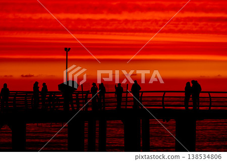 Silhouettes of people on pier at sunset over sea with dramatic red sky, group of figures against ocean horizon, evening atmosphere, lifestyle and travel concept with scenic coastal view 138534806