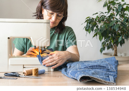 An adult woman repairs and alters denim jeans on a sewing machine at home, focused on an upcycling diy craft project. Reusing things. Environmental awareness. Eco-friendly lifestyle An adult woman repairs and alters denim jeans on a sewing machine at home, focused on an upcycling diy craft project. Reusing things. Environmental awareness. Eco-friendly lifestyle 138534948