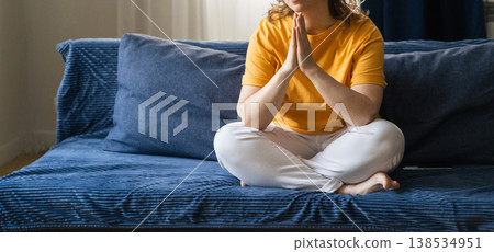 Woman sitting on couch in lotus position, concentrating on meditation with hands in namaste gesture for wellness. Woman practicing yoga meditation techniques Woman sitting on couch in lotus position, concentrating on meditation with hands in namaste gesture for wellness. Woman practicing yoga meditation techniques 138534951