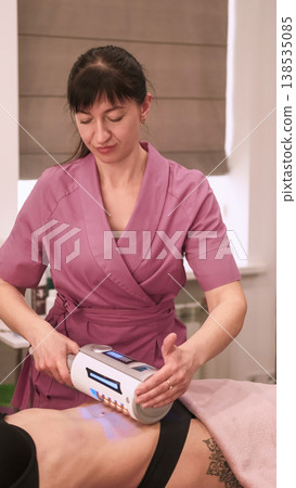 Correction figure. Modern massage. Female beautician specialist using electronic device on woman belly skin for professional endosphere therapy. 138535085