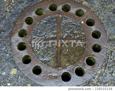 Close-up of a rusty round cast iron manhole cover with drainage holes on wet asphalt. 138535238