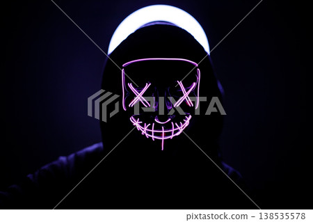Person in Dark Hood Wearing Glowing Neon Purple Mask with X-Shaped Eyes, Atmospheric Portrait with Backlit Halo Effect, Cyberpunk and Mystery Concept Photography in Total Darkness. Person in Dark Hood Wearing Glowing Neon Purple Mask with X-Shaped Eyes, Atmospheric Portrait with Backlit Halo Effect, Cyberpunk and Mystery Concept Photography in Total Darkness. 138535578