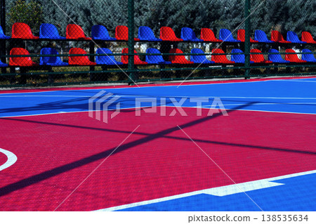 Colorful basketball court with blue and red flooring and empty seating in a sunny outdoor setting Colorful basketball court with blue and red flooring and empty seating in a sunny outdoor setting 138535634