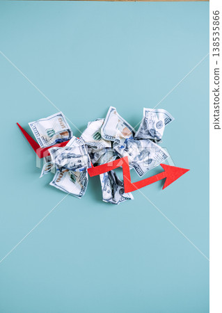 Red arrow points downward over pile of crumpled dollar bills on blue background. Economic recession, deflationary pressure, financial market crash. 138535866
