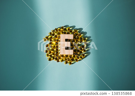 Small wooden letter E stands centered among group of yellow supplement capsules on blue background. Beauty Benefits of Vitamin E, nutricosmetics, glowing skin, skin elasticity. Small wooden letter E stands centered among group of yellow supplement capsules on blue background. Beauty Benefits of Vitamin E, nutricosmetics, glowing skin, skin elasticity. 138535868