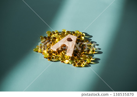 Small wooden letter A stands centered on pile of yellow translucent capsules on blue surface. Respiratory health, Vitamin A deficiency, cellular growth, protective biology. Small wooden letter A stands centered on pile of yellow translucent capsules on blue surface. Respiratory health, Vitamin A deficiency, cellular growth, protective biology. 138535870