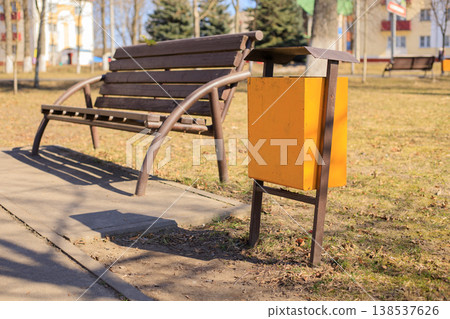 Yellow trash bin in a city park next to a brown bench and a pedestrian path. Yellow trash bin in a city park next to a brown bench and a pedestrian path. 138537626