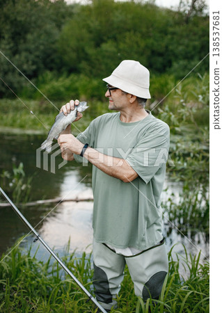 Man with fishing rod near the lake fishing at summer. Fisherman catch a fish. Man wearing khaki clothes and a cap. Man with fishing rod near the lake fishing at summer. Fisherman catch a fish. Man wearing khaki clothes and a cap. 138537681