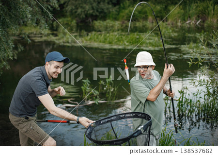 Father and adult son with fishing rods near the lake fishing at summer. Fishermen talking together. Men wearing khaki clothes and a caps. 138537682