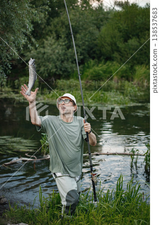 Man with fishing rod near the lake fishing at summer. Fisherman catch a fish. Man wearing khaki clothes and a cap. 138537683