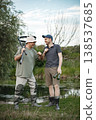 Father and adult son with fishing rods near the lake fishing at summer. Fishermen talking together. Men wearing khaki clothes and a caps. 138537685