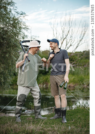 Father and adult son with fishing rods near the lake fishing at summer. Fishermen talking together. Men wearing khaki clothes and a caps. 138537685