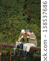 Father and adult son with fishing rods near the lake fishing at summer. Fishermen sitting on a footbridge and drinking a beer. Men wearing khaki clothes and a caps. 138537686