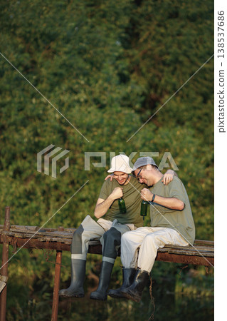Father and adult son with fishing rods near the lake fishing at summer. Fishermen sitting on a footbridge and drinking a beer. Men wearing khaki clothes and a caps. Father and adult son with fishing rods near the lake fishing at summer. Fishermen sitting on a footbridge and drinking a beer. Men wearing khaki clothes and a caps. 138537686