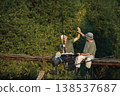 Father and adult son with fishing rods near the lake fishing at summer. Fishermen sitting on a footbridge. Men wearing khaki clothes and a caps. 138537687
