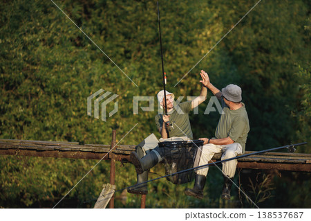 Father and adult son with fishing rods near the lake fishing at summer. Fishermen sitting on a footbridge. Men wearing khaki clothes and a caps. Father and adult son with fishing rods near the lake fishing at summer. Fishermen sitting on a footbridge. Men wearing khaki clothes and a caps. 138537687